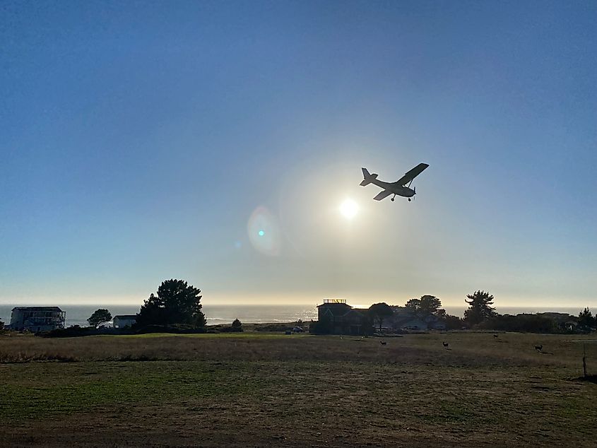 A small charter plane flies over a small golf course, preparing to land against an almost setting sun