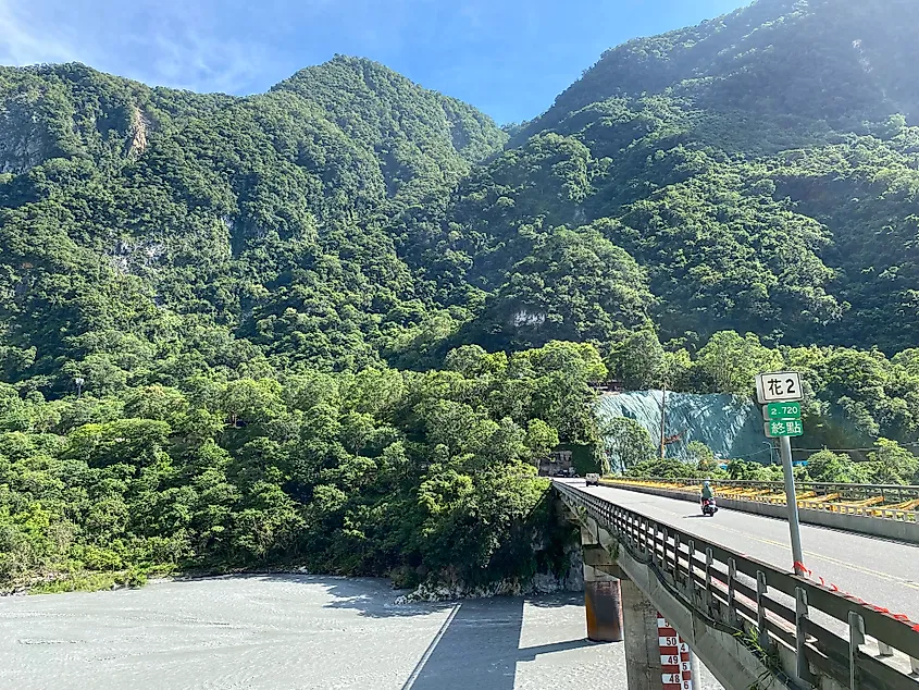 A motorcyclist crossing a river bridge toward steep, lush mountains.
