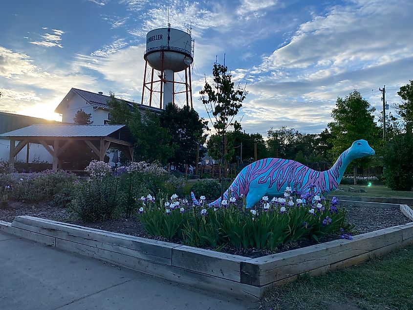 A blue and purple brontosaurus sculpture stands in a pretty garden beneath a small-town water tower.