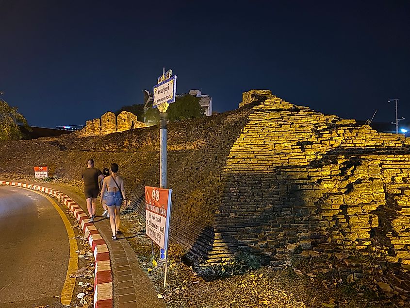 At night, a group of three people walk on a narrow sidewalk beside an ancient stone wall.