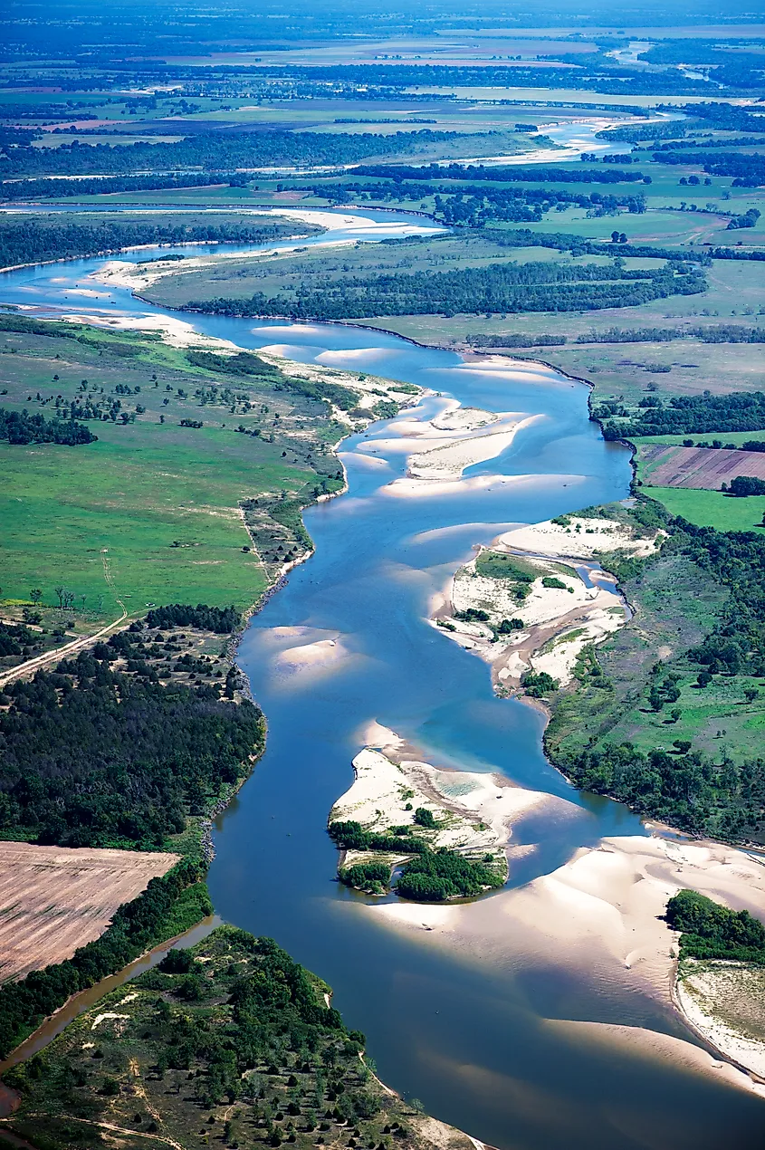 Aerial view of a meandering river snaking through expansive green fields and patches of trees. Sandbanks line the river, creating a peaceful, natural scene.