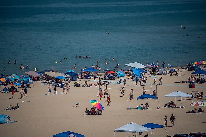 A wide sandy beach in Porter, Indiana, is filled with people swimming, relaxing, and playing under colorful umbrellas and pop-up tents.