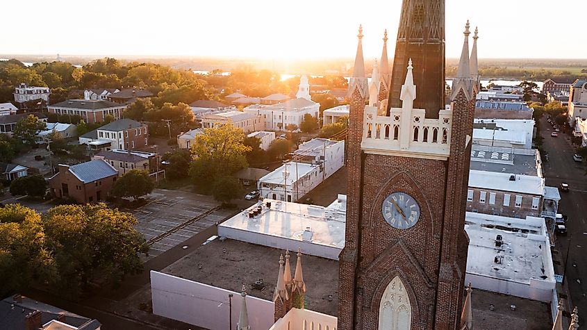Aerial view of downtown Natchez, Mississippi, at sunset, featuring a tall red-brick church with a clock tower and Gothic-style spires. Surrounding the church are historic buildings, tree-lined streets, and the Mississippi River visible in the distance.