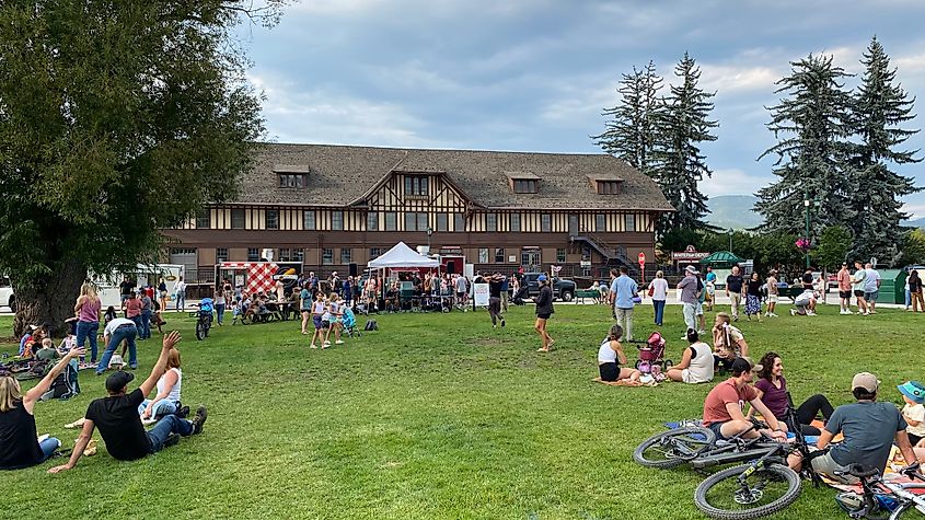 A summer crowd gathers in a park in front of an old train depot.