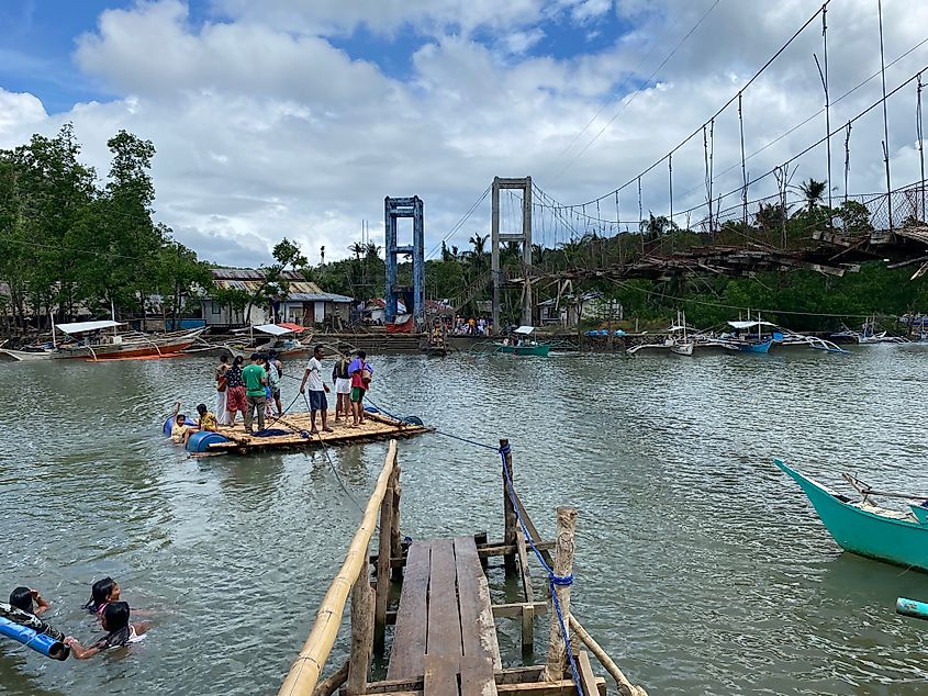 A makeshift wooden raft full of Filipino occupants makes its way across a river next to a washed-out suspension bridge.