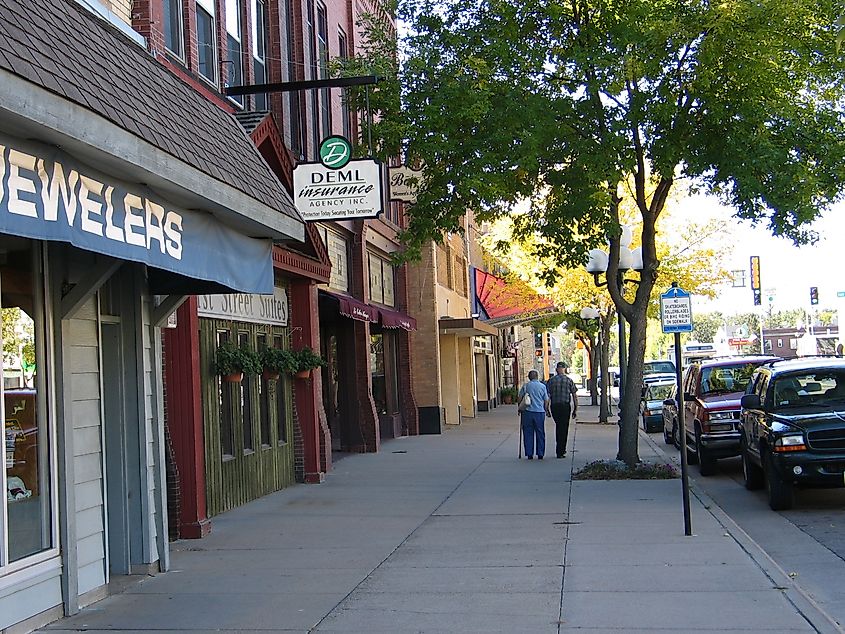 A quaint small-town street with shops, including a jeweler and insurance agency. Two people walk under green trees, exuding a peaceful, welcoming vibe.