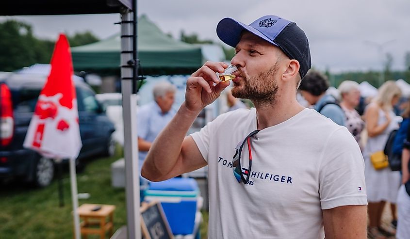 A man in a white t-shirt and blue cap drinks a small glass of alcohol at an outdoor event in limbazi, Latvia.