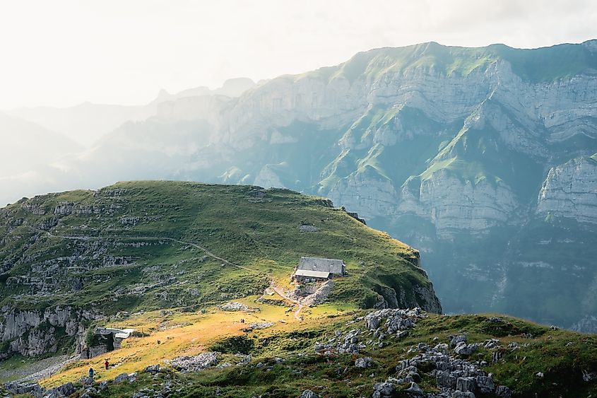 A small hut on a green alpine ridge in Appenzell overlooks massive, sunlit Swiss Alps cliffs.