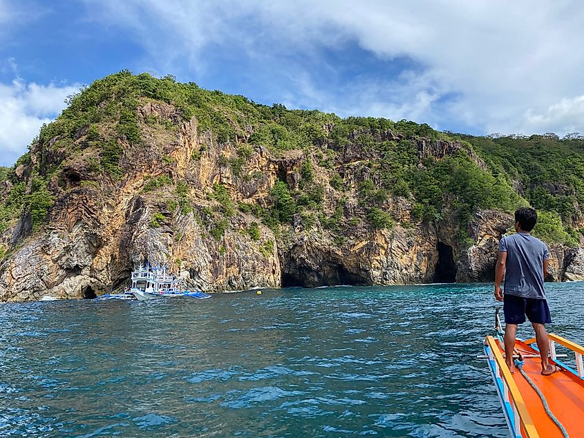 A Filipino man stands at the tip of a tour boat as it approaches a large rocky island during a multi-day island-hopping trip.