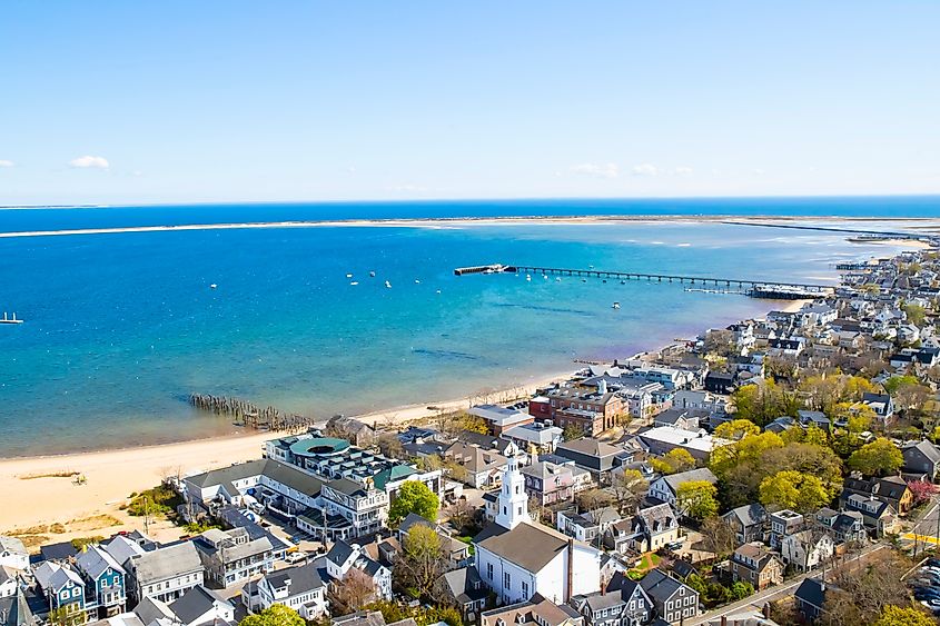 View of the harbor next to the buildings of Provincetown, Massachusetts, with a sandy beach along the shore and a clear blue sky.