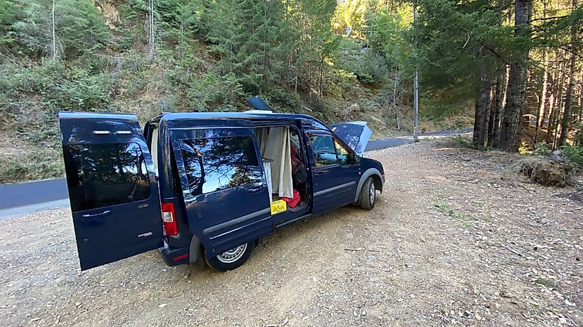 A blue Ford Transit Connect sits on a forested roadside turnout with hood popped.