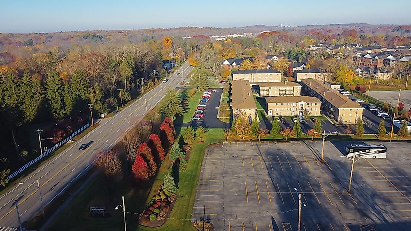 Aerial view of a large vacant parking lot near a multi-story apartment complex along Monroe Avenue in Pittsford, New York, USA, with downtown Rochester and Pinnacle Hill in the background.