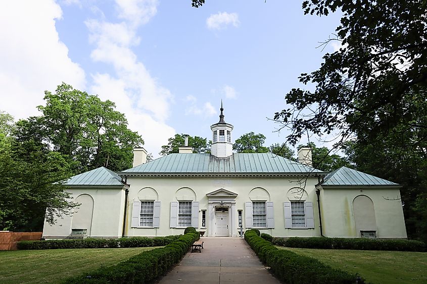 Historic white building with a green roof and cupola, surrounded by lush greenery and clear blue sky. A manicured path leads to the entrance.