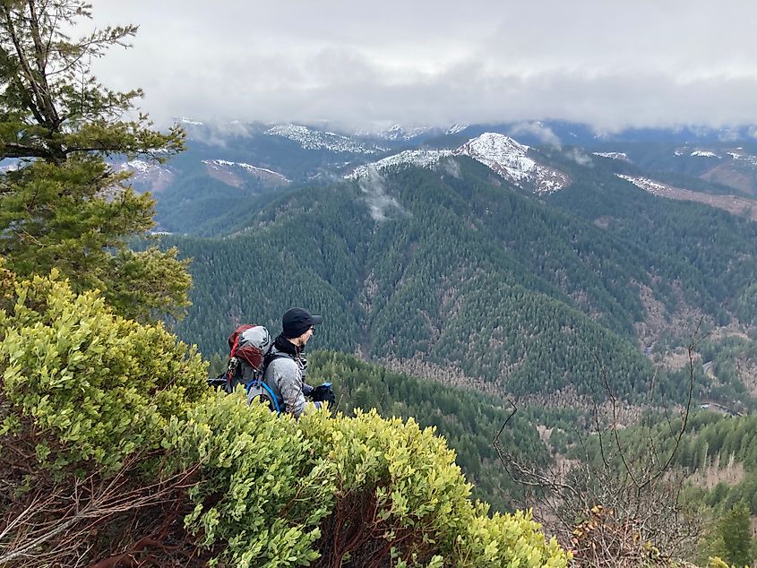 Two hikers, bundled in winter gear, admire a vast, snowy mountain landscape. Dense evergreen forests stretch below under a cloudy sky.