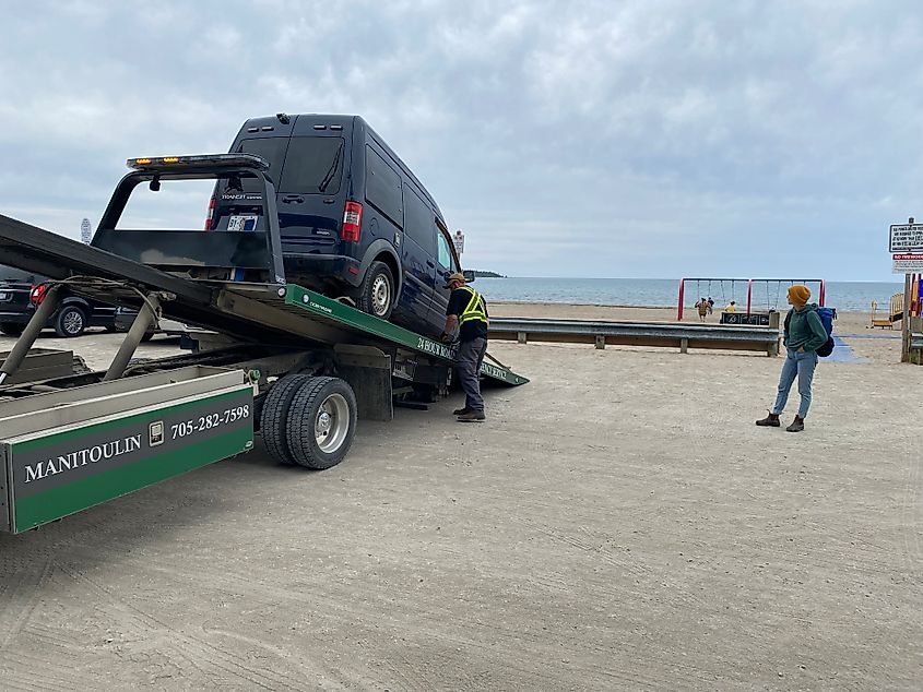 A woman waits beside her blue van that's being hooked up to a tow truck. At least she's at the beach!