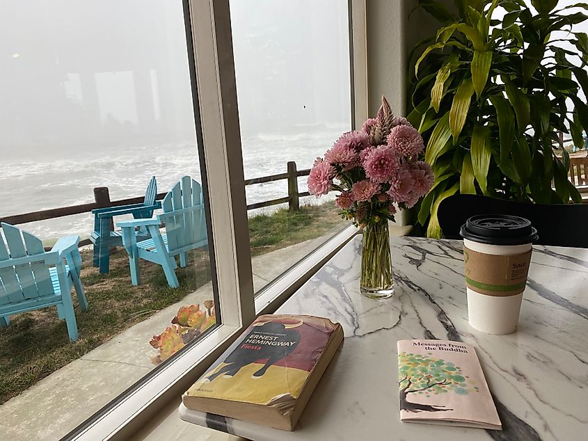 Looking out the window of a quaint coffee shop at a moody ocean. Books and a cup of coffee rest on a white marble table.  