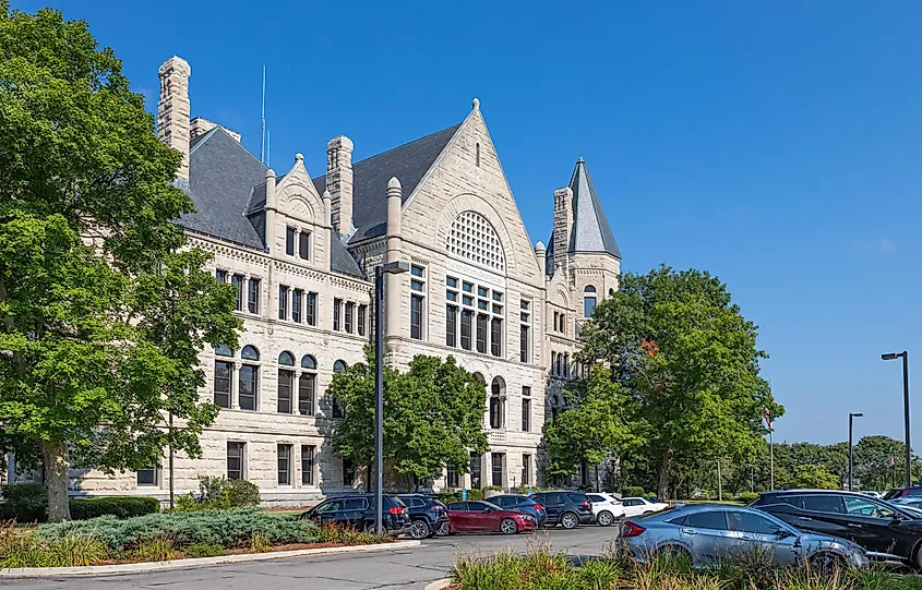 Historic stone building with a slate roof, surrounded by lush green trees, sits under a clear blue sky. Cars are parked in front.