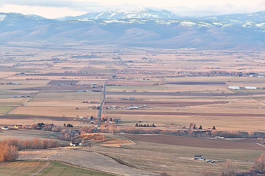 Aerial view of a vast rural landscape with rectangular farm fields in muted browns and greens. Snow-capped mountains loom in the background under a cloudy sky.
