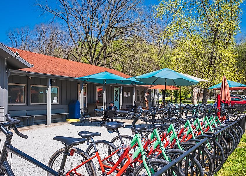 View of the patio of a Midwestern restaurant and bike shop on a nice sunny day in Rocheport, Missouri. In the foreground, there is a row of bicycles, with the café building, umbrellas, and several people visible behind them.