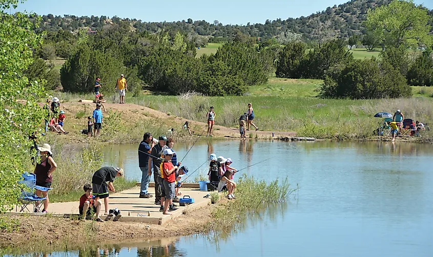 People of all ages fishing along the edge of a small pond in Walsenburg, Colorado, with grassy banks, trees, and low hills in the background under a clear sky.