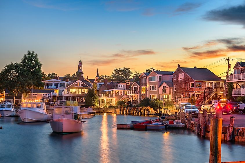 Harbor in Rockport, Massachusetts at sunset with small boats on calm water and colorful coastal houses glowing with evening lights.