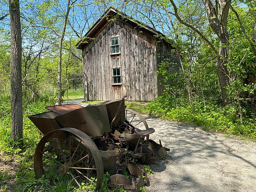 Old rusty farm machinery sits beside a forest trail and an old wooden cabin.