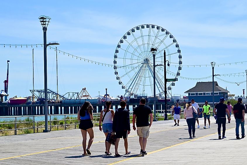 People walking in groups along the Atlantic City Boardwalk in New Jersey, with a Ferris wheel visible in the background and a lively atmosphere under a bright sky.