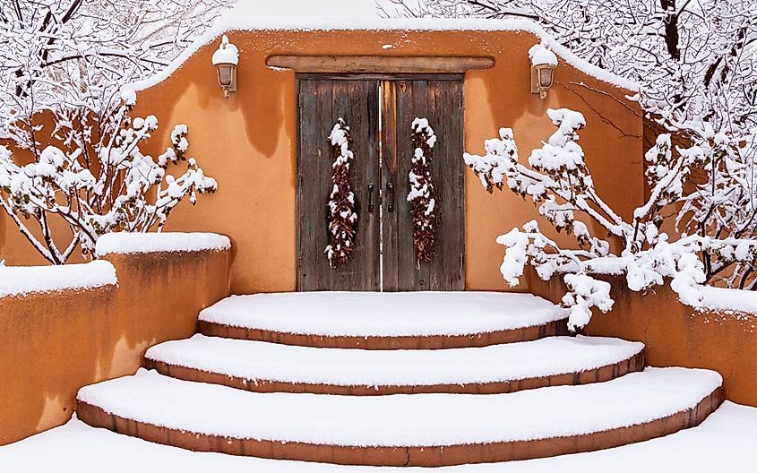 Winter scene of snow-covered adobe wall with rustic wood doors and chile ristras in Santa Fe, New Mexico.