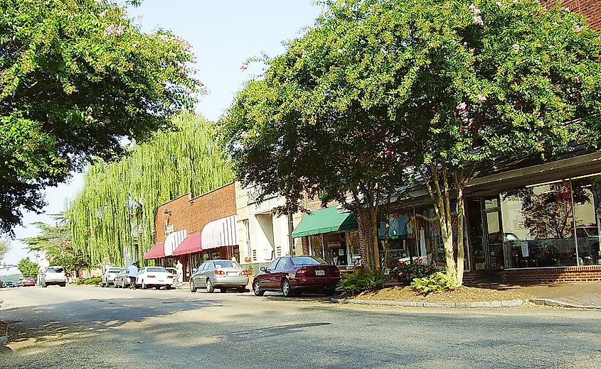 Downtown Smithfield, Virginia, featuring a charming street lined with brick buildings, storefronts with colorful awnings, parked cars, and lush trees providing shade along the sidewalk.