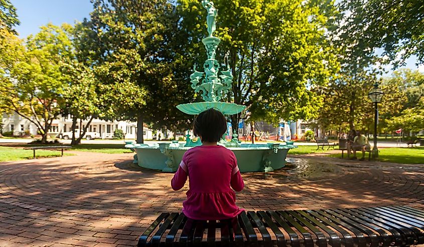 A little girl wearing pink casual clothing is sitting alone on a metal bench in the Fountain Park of Chestertown, Maryland.