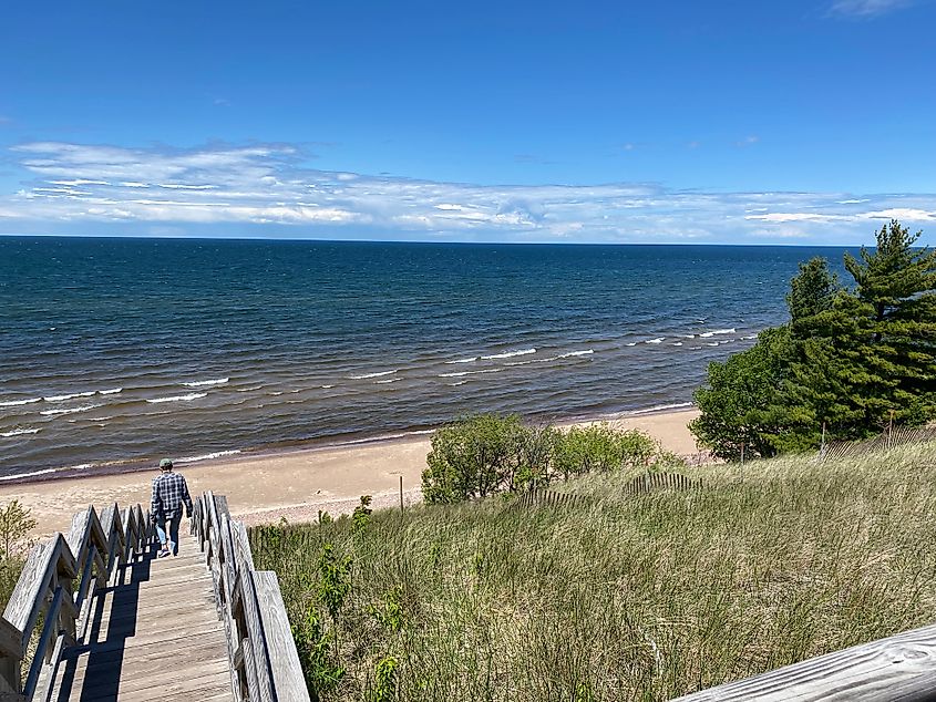 A woman walks down a long wooden staircase toward a sunny and Sandy Lake Superior beach