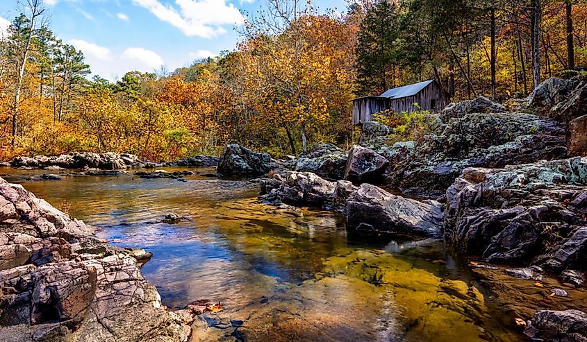 A serene autumn scene with a clear river reflecting the sky, surrounded by rocky banks and vibrant fall foliage. A rustic cabin sits nestled among trees.