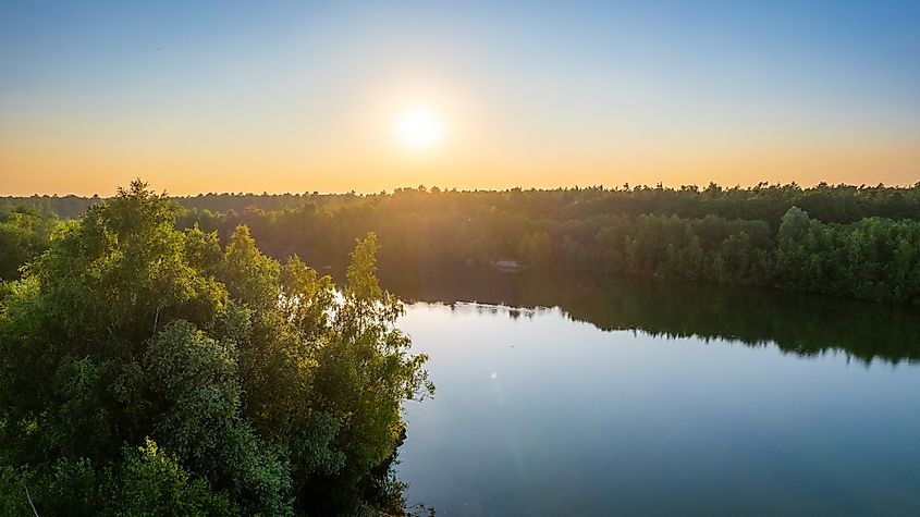 A serene sunset over a still lake, with warm golden and orange hues reflecting on the calm water, surrounded by a dense, lush forest silhouetted against the fading light.