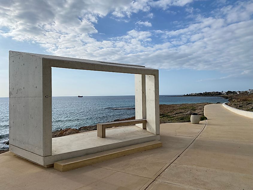 An offshore ship seen through a rectangular stone sculpture situated on a paved promenade.