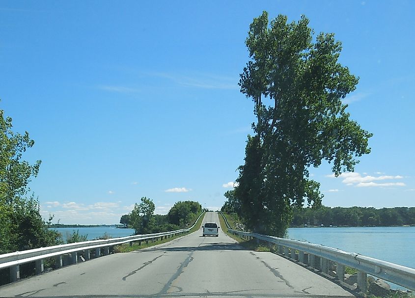 A view of the causeway leading to Johnson's Island, Ohio, with a vehicle driving towards the island on a sunny day. The road is flanked by water on both sides, with trees and greenery in the background.