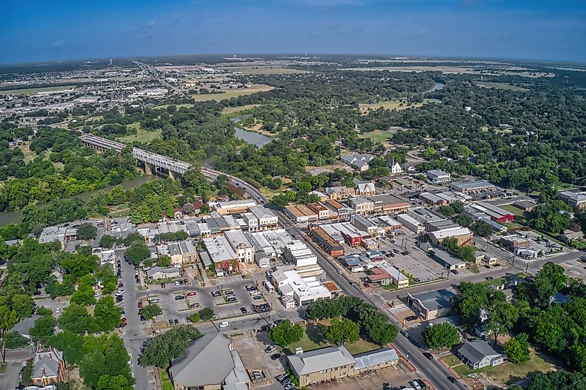 Aerial view of the town of Bastrop, Texas, showcasing a mix of historic buildings, tree-lined streets, and the nearby Colorado River winding through the landscape.