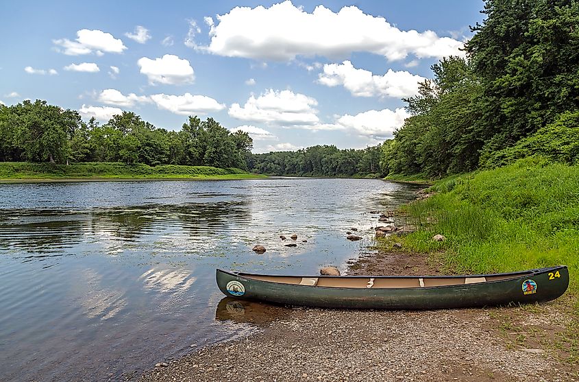 A green canoe rests on a pebbled riverbank, surrounded by lush greenery. The calm river reflects a blue sky with puffy white clouds, evoking tranquility.