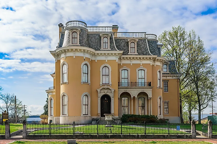 Elegant Victorian-style mansion with ornate arched windows, a mansard roof, and a wrought iron fence, set against a partly cloudy blue sky.