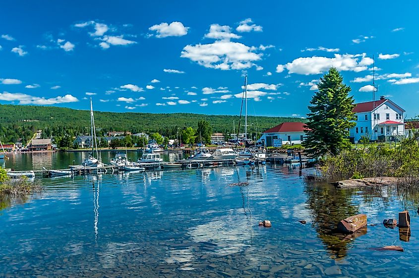 Scenic lakeside view with clear blue skies, white clouds, and sailboats docked at a marina. Lush green trees and charming buildings line the shore.