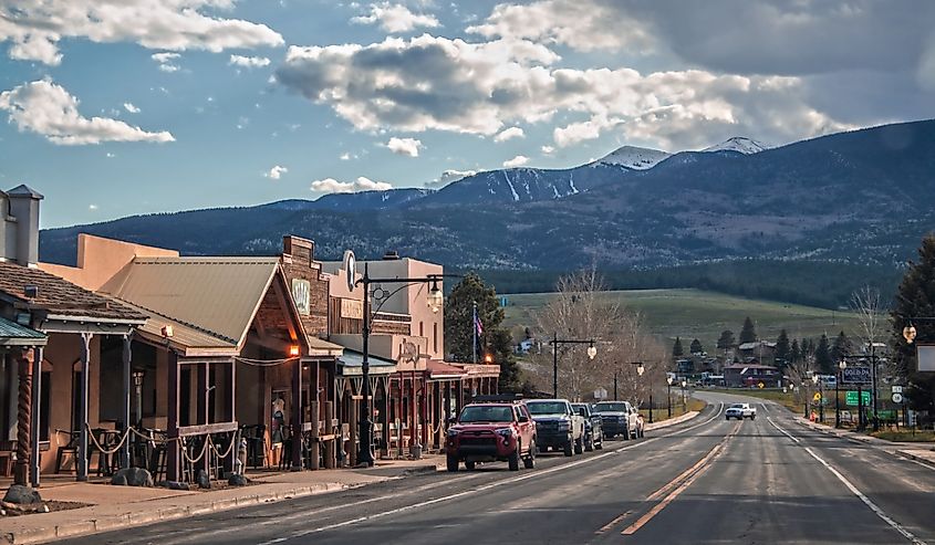 Small western style tourist town near Angel Fire ski resort in New Mexico with cars parked on main drag through town and mountains in background at dusk