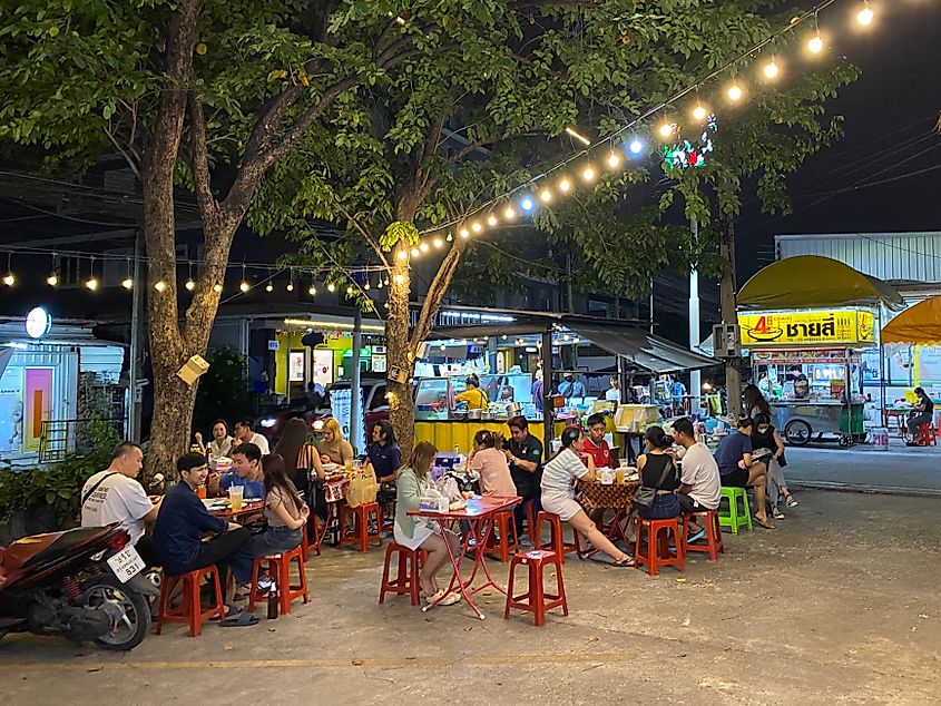 Groups of patrons sit at red tables in the parking lot of a pop-up food vendor. Strung lights illuminate the Bangkok night.