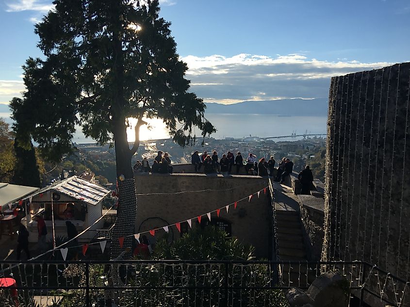 A group gathers atop a hilltop castle as the sun begins to set over the waterfront city below.