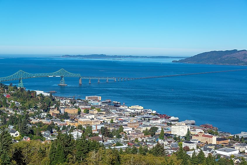 Coastal cityscape with a large bridge spanning blue water. Buildings cluster in the foreground, flanked by lush green hills under a clear blue sky. Peaceful vibe.