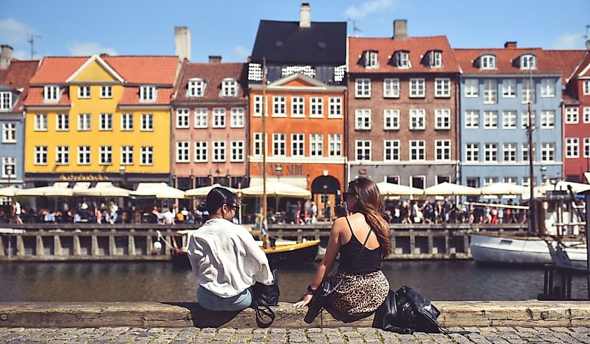 Two women sit on background on embankment of canal with colorful buildings in Copenhagen