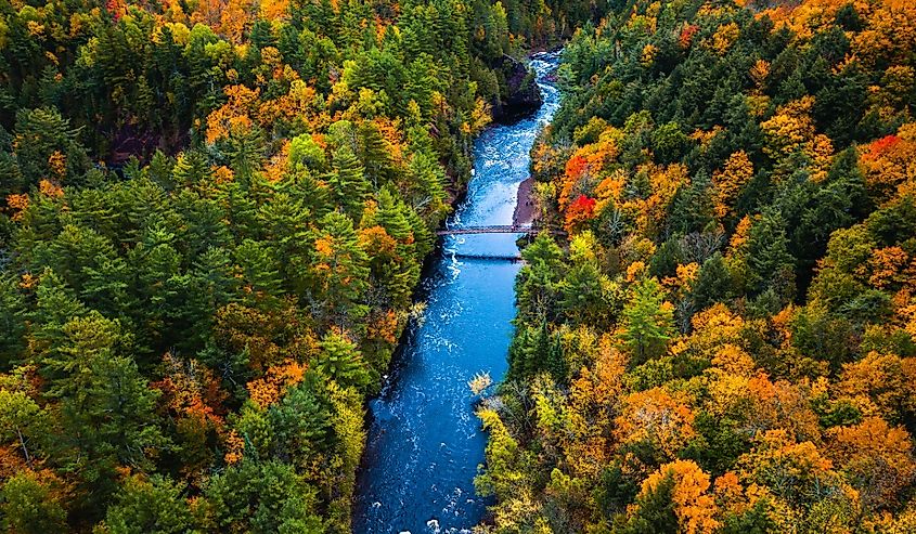 Beautiful travel aerial of a pedestrian foot bridge crossing the bright blue water of the Bad River at Copper Falls with colorful fall foliage lining the river banks in autumn in Mellen, Wisconsin.