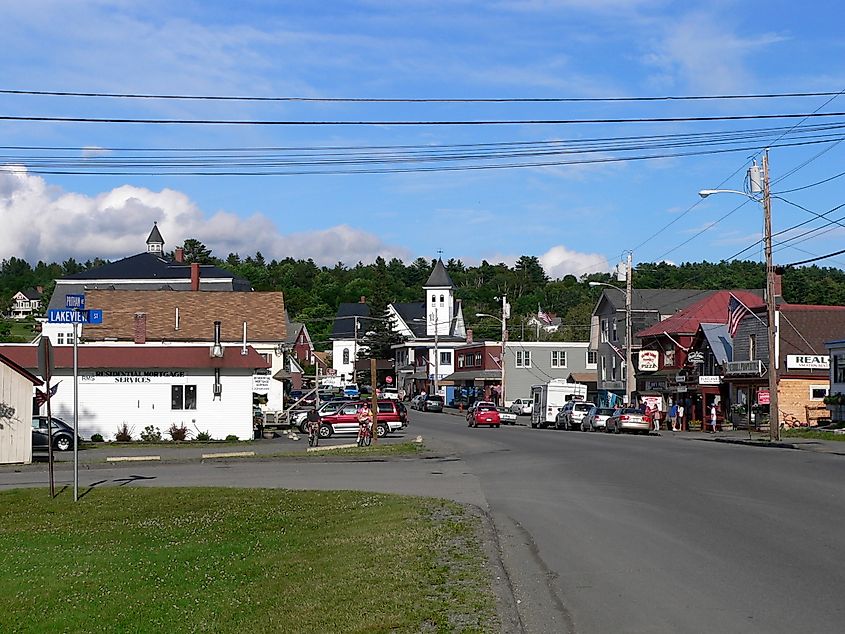 View of Greenville, Maine's main street, featuring small businesses and local traffic, with hills and a partly cloudy sky in the background.