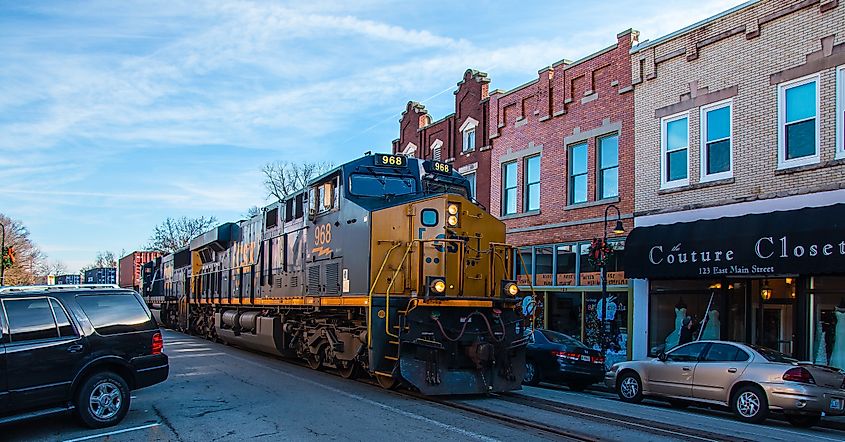 A large CSX freight train passes through the center of La Grange, Kentucky, running down Main Street alongside parked cars and storefronts. 