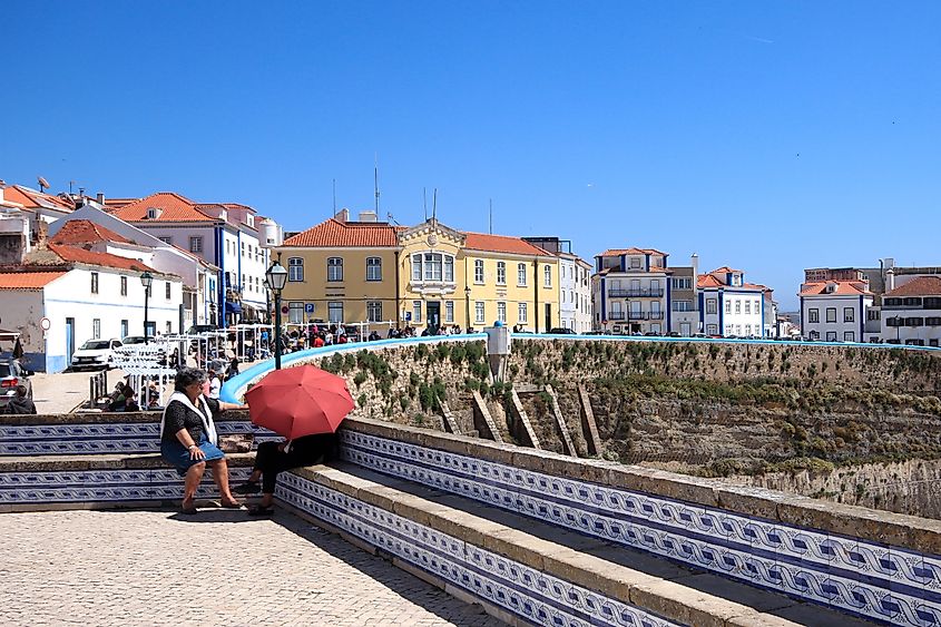 A view of Ericeira village in Portugal, featuring whitewashed buildings with red-tiled roofs perched on cliffs overlooking the Atlantic Ocean, with waves crashing below and a clear blue sky above.