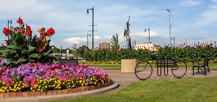 A vibrant garden features red and pink flowers encircled by a brick border. A bike rack and bench sit on a walkway under a blue sky.