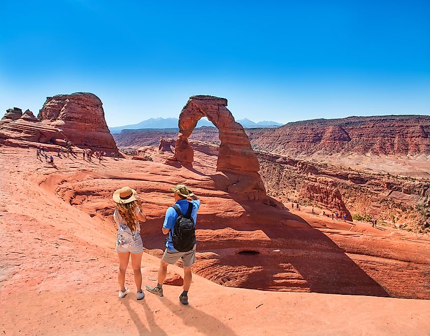 A couple standing on top of a mountain, looking out at the view of Delicate Arch in Arches National Park, Moab, Utah.